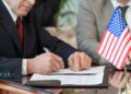 Mature American delegate in formalwear signing contract of business partnership with foreign partner while sitting by table in boardroom