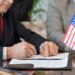Mature American delegate in formalwear signing contract of business partnership with foreign partner while sitting by table in boardroom
